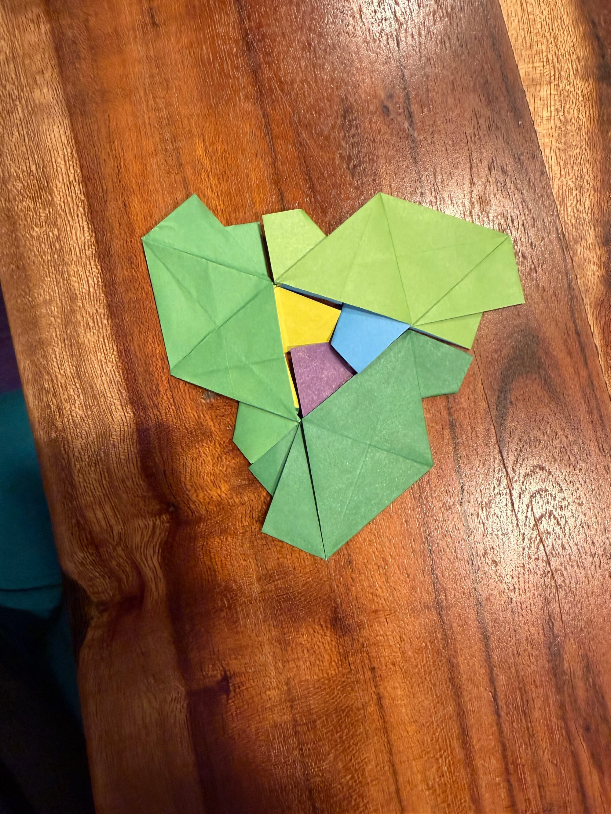 three aperiodic turtle origami peices with all three heads in the center forming a triangle. the tiles are all green with one leg a different color. one is blue, one is purple, and one is yellow. they are on a wooden table. 