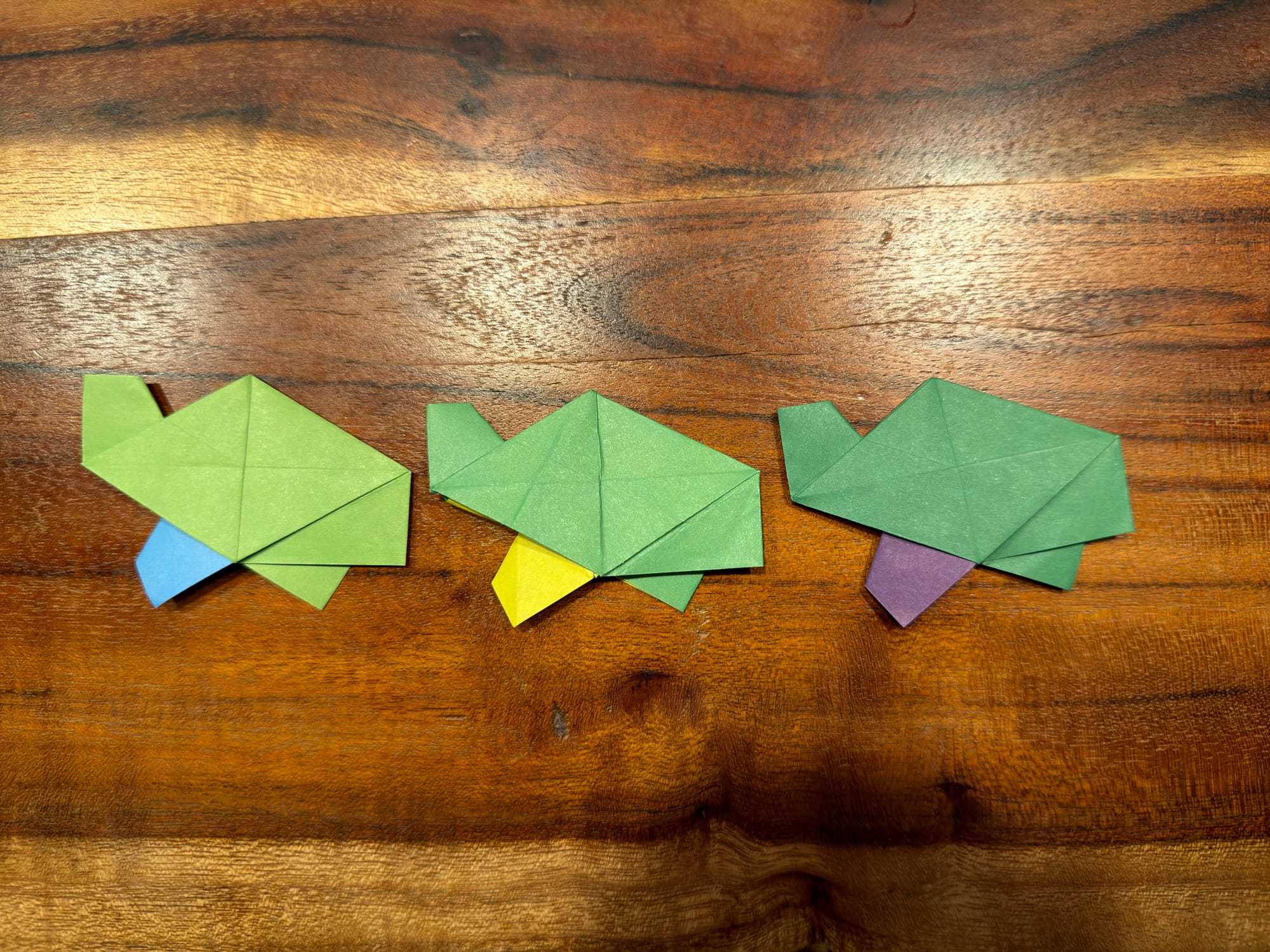 three aperiodic turtle origami peices with all in a row. the tiles are all green with one leg a different color. one is blue, one is purple, and one is yellow. they are on a wooden table. 