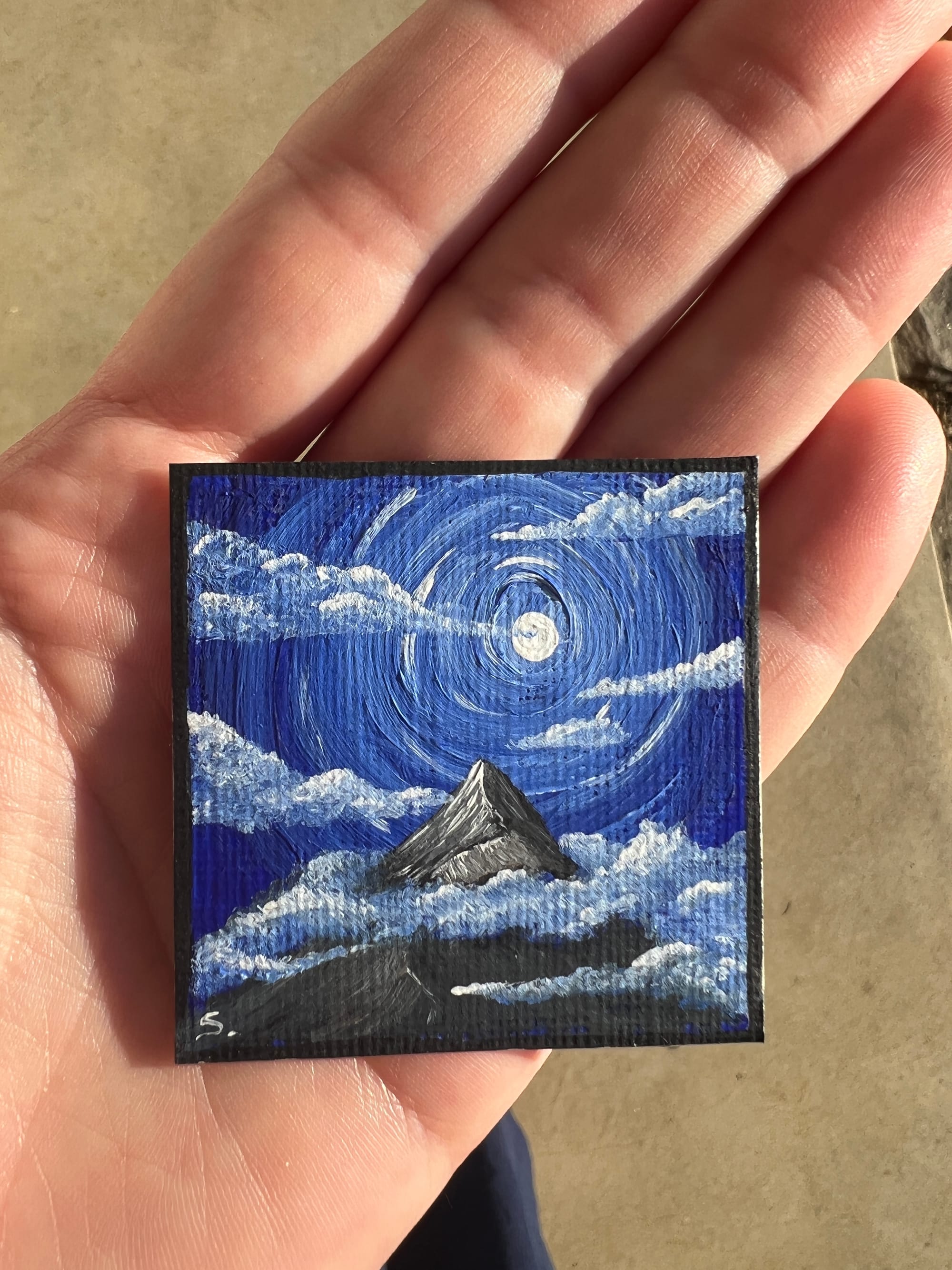 a hand holding a small 2in painting of a mountain with clouds with a moon and blue sky.